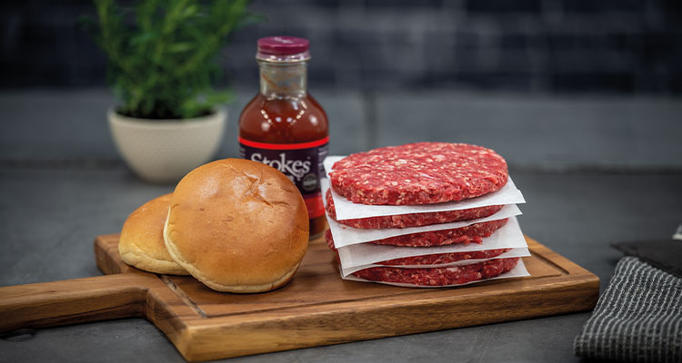 five fresh uncooked burgers on a wooden board with two buns & a bottle of sauce