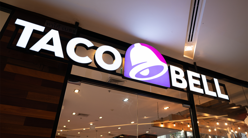 Taco Bell restaurant storefront with illuminated logo signage above glass entrance and warm interior lighting visible inside
