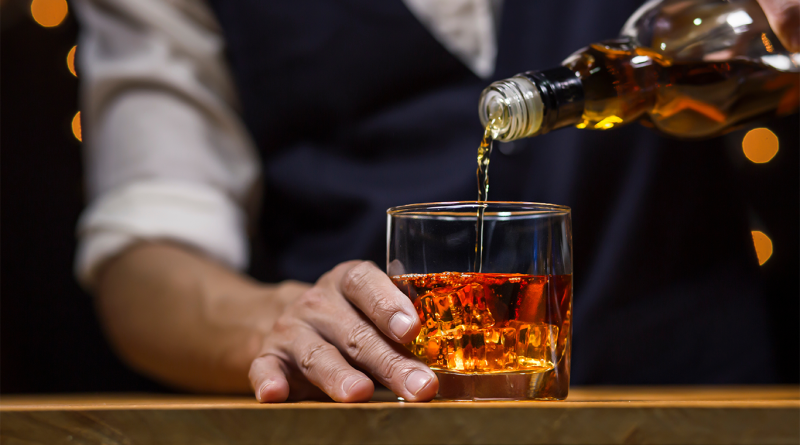 Whiskey being poured into a glass over ice by a bartender, with warm ambient lighting and a blurred background.