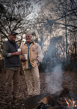 two men standing in a wooded area near a campfire