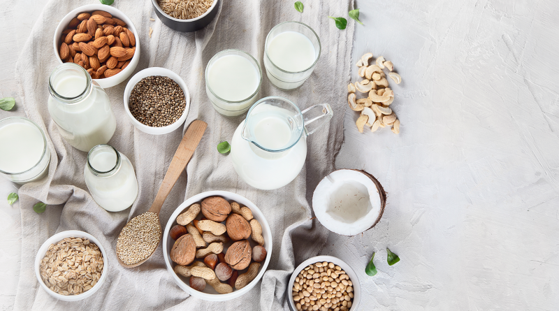 Assorted plant-based milk alternatives including oat, almond, soy, coconut and nut-based beverages arranged on a neutral fabric surface with raw ingredients in bowls and glass containers.