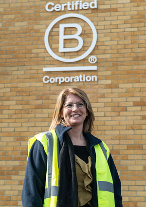  woman standing in front of a light brown brick wall. Behind her, there is a large, white "Certified B Corporation" logo