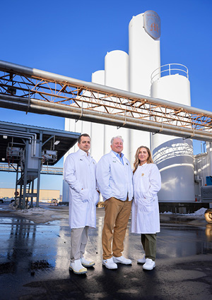 three individuals in white lab coats standing in front of large cylindrical storage tanks