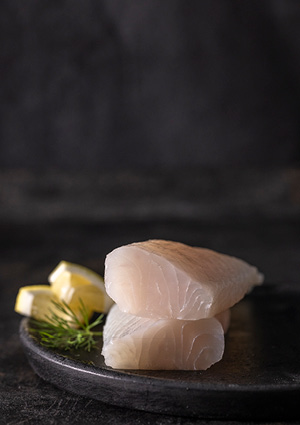 two raw cod fillets stacked on a dark plate, accompanied by lemon wedges and fresh dill