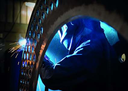 a worker wearing a protective welding helmet and dark workwear is actively welding a heavy structural metal component