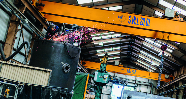 a worker grinding a large metal tank beneath heavy yellow overhead cranes