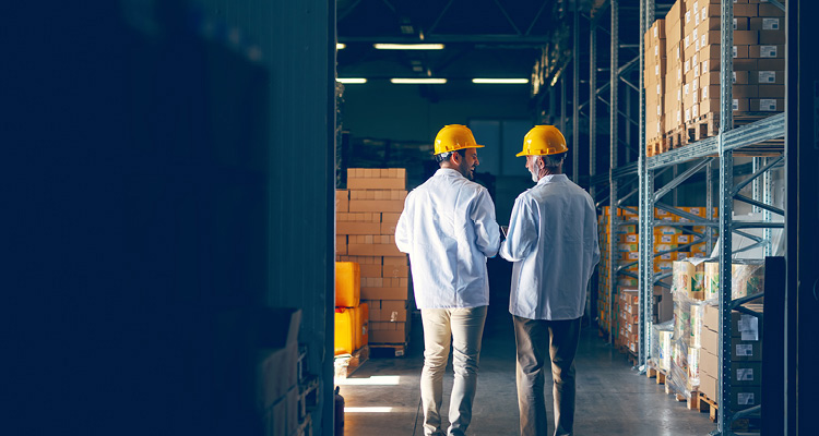 two warehouse workers in yellow hard hats and white shirts walking down an aisle lined with storage racks and stacked cardboard boxes