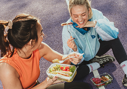 two people sitting outdoors on a paved ground, sharing a healthy meal from reusable or clear takeaway containers