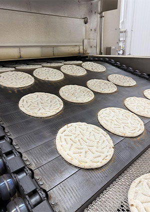 raw or partially baked flatbread or pizza dough rounds moving along an industrial conveyor belt into a commercial tunnel oven