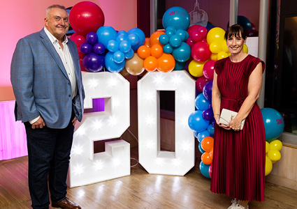 event photo celebrating a 20th anniversary, featuring a man and a woman posing next to giant, illuminated "20" light-up numbers