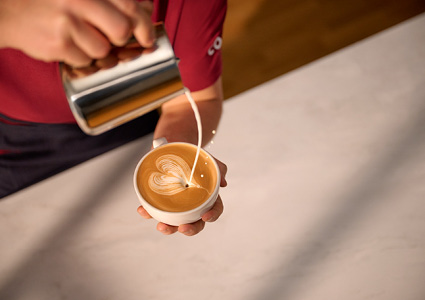A barista is pouring steamed milk from a metal jug into a ceramic cup to create latte art, featuring a classic heart design on top of a frothy cappuccino