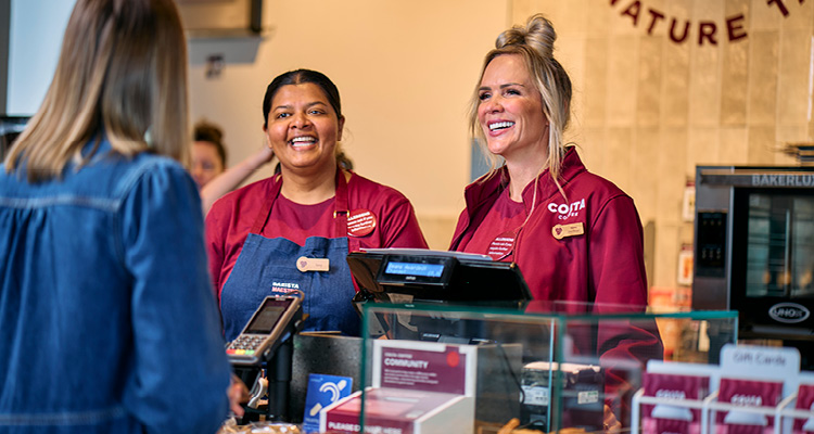 smiling Costa Coffee team members in burgundy uniforms attending to a customer at a service counter equipped with a card payment terminal