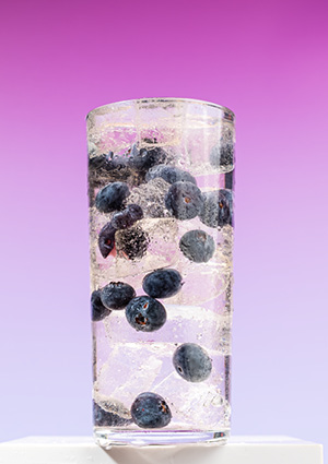 a tall, clear glass filled with ice cubes and whole fresh blueberries suspended in a cold, sparkling beverage