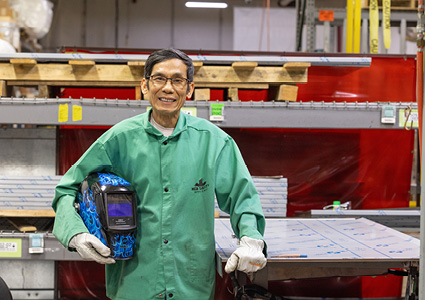 a person wearing safety gear and a green work jacket, holding a blue welding helmet inside an industrial workshop