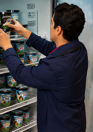 a person in a dark blue jacket stocking a commercial glass-door freezer filled with various tubs of Ben & Jerry's ice cream