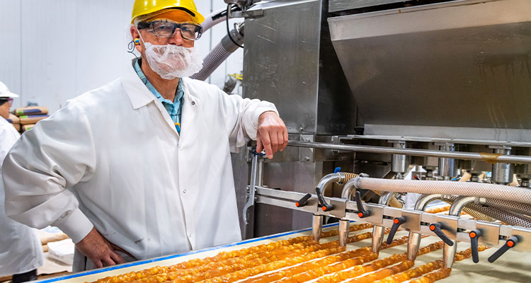 Bakery worker wearing protective clothing and a hard hat beside an industrial production line filled with baked goods.