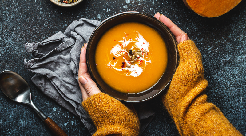 Alt text Hands in mustard-yellow sweaters holding a bowl of creamy pumpkin soup topped with cream and seeds on a dark rustic table.