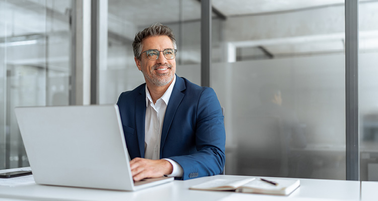a middle-aged businessman wearing glasses and a blue suit while working on a laptop at an office desk