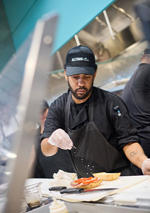 a kitchen employee wearing a black uniform and an Alltown Fresh cap while preparing food at a counter
