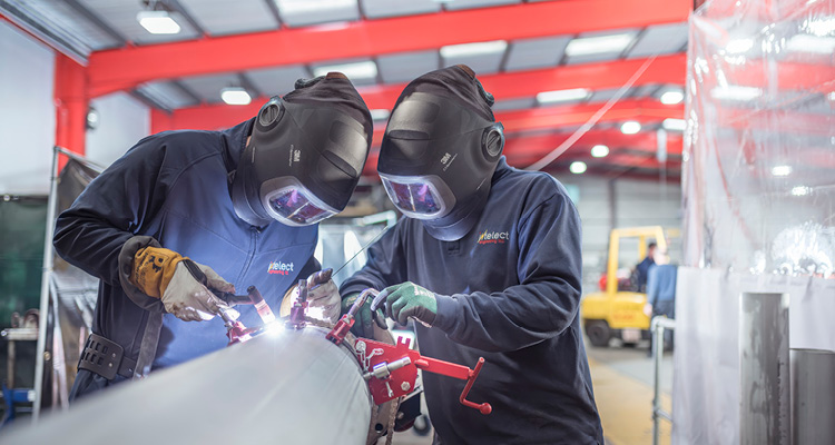 two people wearing protective welding helmets and gear while working together to weld a large metal pipe in an industrial workshop