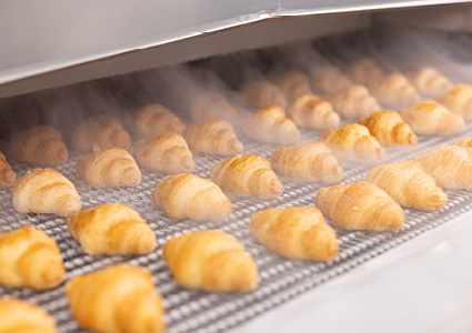 golden brown croissants on a wire conveyor belt inside an industrial bakery oven with steam visible